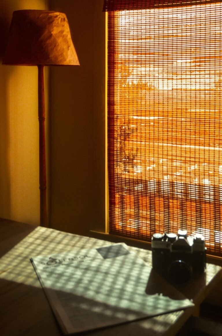 Dark room interior with light on wooden table with newspaper and vintage photo camera near lamp and window covered with jalousie